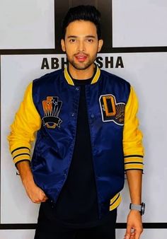 a man standing in front of a white wall wearing a blue and yellow baseball jacket