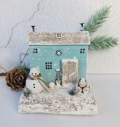 a small house with two snowmen in front of it and pine cones on the side