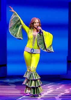 a woman in a yellow outfit on stage with her arms out and hands up to the side