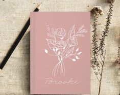 a pink notebook sitting on top of a table next to dried flowers and a pen