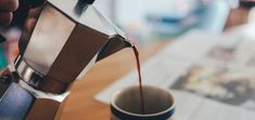 a person pours coffee into a cup on top of a wooden table next to a newspaper