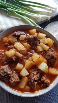 a bowl filled with meat and potatoes on top of a blue table cloth next to bread