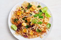 a white plate topped with noodles and veggies next to lime wedges on top of a wooden table