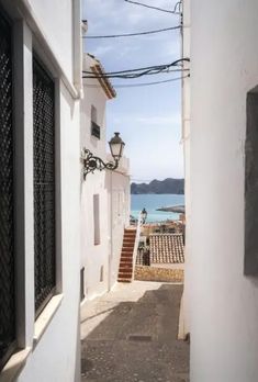 an alley way leading to the beach with white buildings and blue water in the background