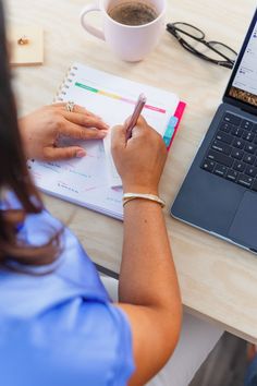 a woman sitting at a desk with a laptop and notebook in front of her, writing on a notepad