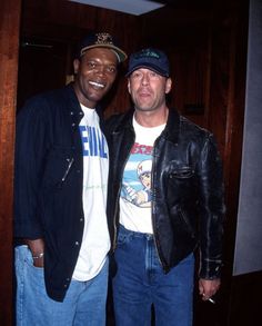 two men standing next to each other in front of a wooden door wearing jackets and jeans