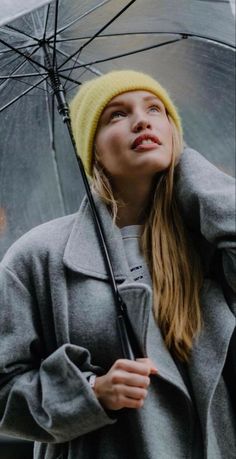 a woman standing under an umbrella in the rain wearing a gray coat and yellow knitted hat