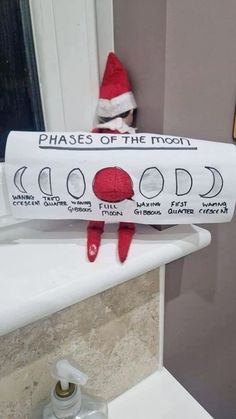 an elf is sitting on the edge of a bathroom sink holding a sign that says phases of the moon