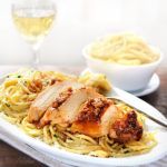 a white plate topped with chicken and pasta next to a glass of wine on top of a table