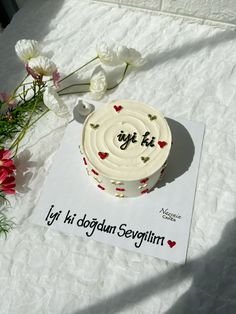 a white cake sitting on top of a table next to flowers and a piece of paper