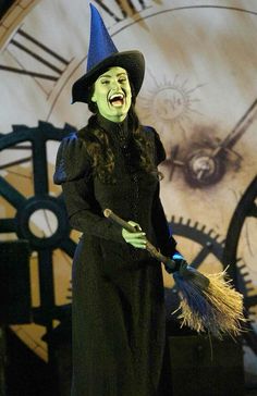 a woman dressed in green and black holding a broom with her mouth open while standing next to a clock