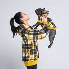 a woman holding a small dog wearing a yellow and black checkered shirt while standing in front of a white wall