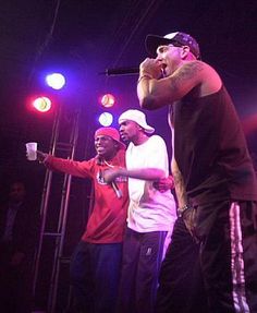 three men standing on stage with one holding a microphone and the other pointing at something