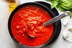 a bowl of tomato sauce with a spoon in it next to garlic and basil leaves