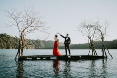 a man and woman standing on top of a wooden dock next to trees in the water