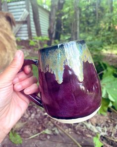 a hand holding a purple and green coffee mug in front of trees with leaves on the ground