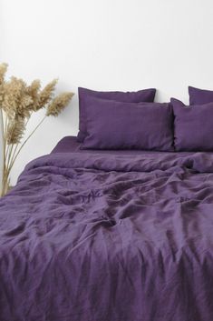 a bed with purple sheets and pillows on it next to a vase with dried flowers