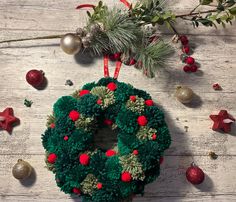 a green christmas wreath with red pom - poms and ornaments on a wooden table