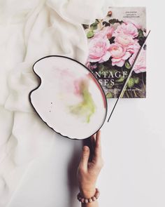 a person holding up a plate with flowers on it next to a book and white sheets