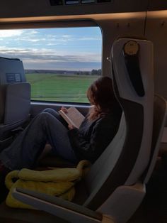 a woman sitting in an airplane looking out the window while reading a book on her lap