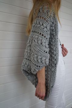 a woman standing against a wall wearing a gray crochet sweater and white pants