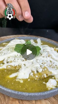 a bowl filled with green food on top of a wooden table next to a person's hand