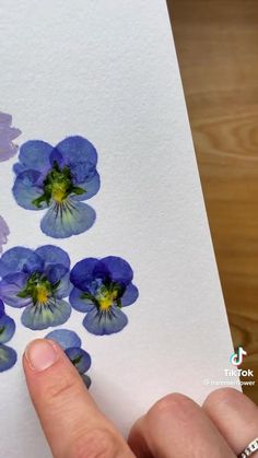 a hand is pointing at some blue flowers on a piece of paper that has been painted with watercolors