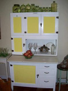 a white cabinet with yellow doors and glassware on it's top, next to a small table