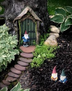 two gnomes are sitting in the garden next to a tree and stone path that leads up to a small house