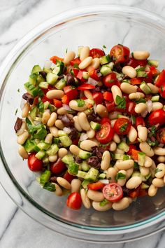 a glass bowl filled with beans, tomatoes and cucumber on top of a marble counter