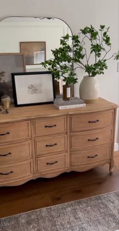 a wooden dresser topped with a mirror and vase filled with green plants next to a framed photograph