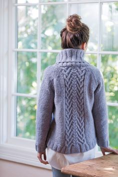 a woman standing in front of a window with her back turned to the camera, wearing a gray cabled sweater