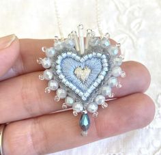 a hand holding a heart shaped pendant with pearls and beads on it's fingers