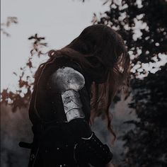 a woman with red hair and armor standing in front of trees, looking down at the ground