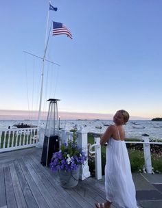 a woman in a white dress standing on a porch next to an american flag and flowers