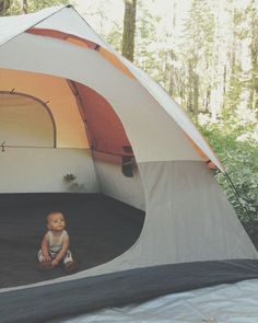 a baby sitting in front of a tent with the door open and trees in the background