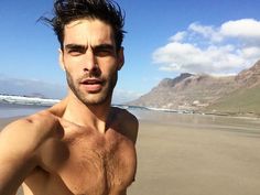 a man standing on top of a sandy beach next to the ocean with mountains in the background