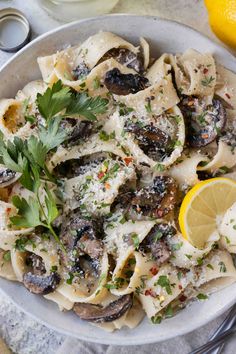 a white bowl filled with pasta covered in mushrooms and parsley next to lemon wedges