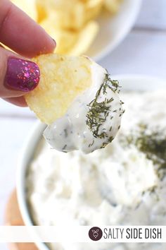 a hand holding a piece of food over a bowl of dip with chips in the background