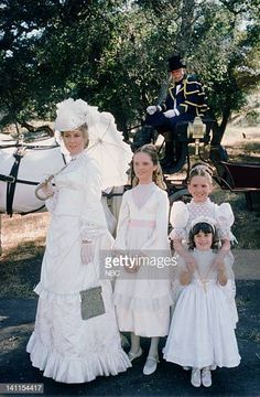 three girls in white dresses standing next to a horse and carriage with two men on horseback behind them