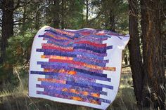 a quilt hanging from the side of a tree in a forest with grass and trees