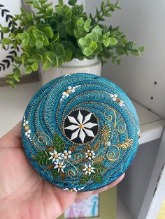 a hand holding a blue beaded rock with white flowers on it and green plants in the background