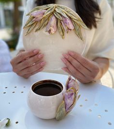 a woman holding a white vase with flowers on it next to a cup of coffee