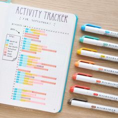 an open notebook with markers and pencils next to it on a wooden table top