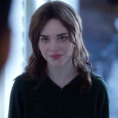 a woman with long brown hair wearing a black shirt and looking at the camera while standing in front of a window