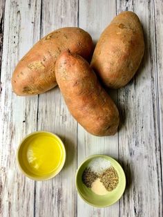 three sweet potatoes next to two small bowls with mustard and seasoning in them on a white wooden surface