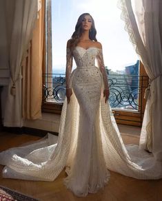 a woman standing in front of a window wearing a wedding dress with long sleeves and an off the shoulder train