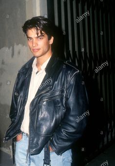 a young man wearing a leather jacket and jeans standing in front of a fence - stock image