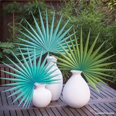 three white vases sitting on top of a wooden table next to each other with green leaves in them