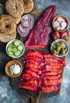 an assortment of meats and vegetables on a cutting board next to bagels, onion rings, pickles, cucumbers